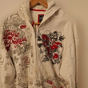 Vit hoodie med tryck från Ecko Unltd. - Säljer en vit hoodie från Ecko Unltd. med dragkedja och stora grafiska tryck i rött, svart och grått över hela tröjan. Hoodien har långa ärmar och ribbade muddar. Perfekt för dig som gillar streetwear och unika detaljer.