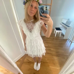 Vit spetsklänning med volanger - Superfin vit kortklänning i spets med volangdetaljer och v-ringning. Klänningen har korta ärmar och flera lager volanger som ger en romantisk känsla. Perfekt till sommarens festligheter eller studentfirande.
