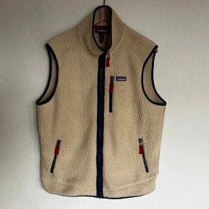 Patagonia fleeceväst  - Säljer en beige fleeceväst från Patagonia med röda detaljer och marinblå dragkedjor. Västen har hög krage, tre fickor med dragkedja och klassisk logga på bröstet. Perfekt för lager-på-lager och enkel att matcha.