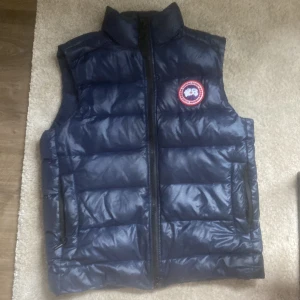 Blå dunväst från Canada Goose - Säljer en blå dunväst från Canada Goose med hög krage och dragkedja framtill. Västen har ett klassiskt märke på bröstet och två fickor med dragkedja. Perfekt för lager-på-lager under kyliga dagar.