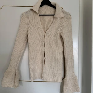 Beige stickad kofta med utsvängda ärmar - Säljer en beige stickad kofta med stor krage och v-ringning. Tröjan har långa, utsvängda ärmar och är i ett mjukt material. Perfekt för lager-på-lager och en stilren look.