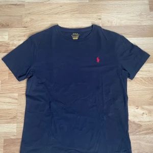 Svart t-shirt från Polo Ralph Lauren - Klassisk svart t-shirt från Polo Ralph Lauren med röd broderad logga på bröstet. Nytt skick, inga defekter eller fläckar. T-shirten har rund halsringning och är tillverkad i mjuk bomull. Perfekt för en stilren och enkel look. Storlek S. 