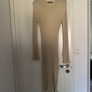 Beige ribbad långklänning - Beige ribbad långklänning med lång ärm och rund halsringning. Klänningen har en tight passform och är tillverkad i ett stretchigt stickat material som sitter snyggt på kroppen. Ärmarna har slits och den vänstra sidan har slits nertill. Ej använd