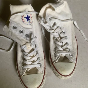 Vita Converse All Star höga sneakers - Klassiska vita Converse All Star high-top sneakers med snörning och rund tå. Skorna har vit canvas, röd och blå rand längs sulan och Converse-logga på sidan. Perfekta för en avslappnad och tidlös stil.