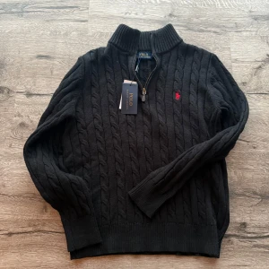 Polo Ralph Lauren stickad tröja - Kabelstickad svart tröja från Polo Ralph Lauren med halv dragkedja och hög krage. Klassisk röd logga broderad på bröstet. Tröjan har långa ärmar och ribbade muddar för extra komfort. Helt ny aldrig använd. Skicka pm vid minsta fundering