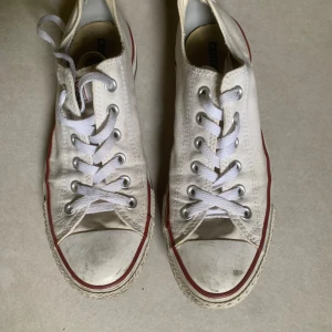 Vita Converse All Star sneakers - Klassiska vita Converse All Star sneakers med lågt skaft och vit gummisula. Skorna har snörning och röda detaljer längs sulan. Perfekta för en avslappnad och tidlös stil.