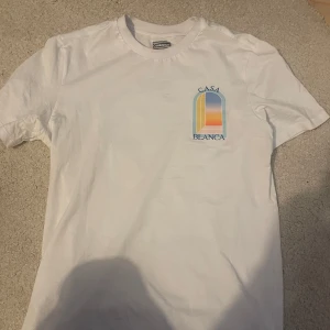 Vit t-shirt från Casablanca med färgglatt tryck - Vit t-shirt från Casablanca med färgglatt grafiskt tryck och texten 'Casa Blanca' på bröstet och ryggen. Klassisk rund hals och normal passform. Perfekt för dig som gillar stilrena plagg med en twist. Aldrig använd bara testat