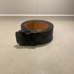 Svart LV bälte - Säljer ett svart bälte från Louis Vuitton med klassiskt rutigt mönster och svart LV-spänne. Bältet har en slät insida i brunt skinn och är perfekt för att lyfta din outfit med en exklusiv touch.