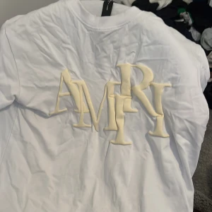 Vit t-shirt från Amiri med 3D-logga - Säljer en vit t-shirt från Amiri med stor, upphöjd 3D-logga framtill i beige. T-shirten har klassisk passform och rund halsringning. Perfekt för dig som gillar streetwear och exklusiva märken.