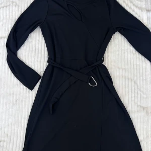 Svart klänning med bälte från LC Waikiki - Snygg svart klänning från LC Waikiki med lång ärm och tillhörande bälte i midjan. Klänningen har en stilren design med en liten öppning vid bröstet perfekt för dig som gillar en klassisk look.