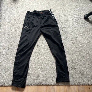 Svarta träningsbyxor från Kappa - Svarta träningsbyxor från Kappa med det klassiska logomärket på vänster ben och längs sidorna. Byxorna har elastisk midja och smal passform, perfekta för sport eller chill. Snygg och enkel design med vita detaljer.