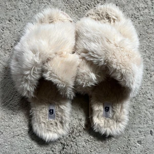 Beiga fluffiga inne tofflor - Supermjuka och fluffiga tofflor i beige färg med korsad design upptill. Perfekta för att mysa runt hemma, med en riktigt gosig känsla tack vare det lurviga materialet.