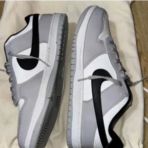 Nike Dunk Low i grått och vitt - Säljer ett par Nike Dunk Low sneakers i grått och vitt med svart swoosh. Skorna har klassisk låg modell, rund tå och platt sula. Perfekta för dig som gillar stilrena och sportiga sneakers.