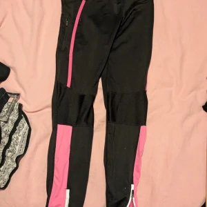 Svarta träningsbyxor med rosa detaljer - Säljer ett par svarta träningsbyxor med rosa detaljer och dragkedjor vid bensluten. Byxorna har meshpaneler för extra ventilation och en dragsko i midjan. Perfekta för dig som vill ha snygga och funktionella träningskläder.