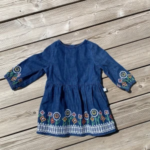 Blå jeansklänning för barn Stella McCartney - Supersöt blå jeansklänning från Stella McCartney med färgglada broderade blommor och staketdetalj nertill. Klänningen har trekvartsärm och knappar i ryggen. 