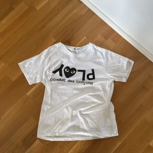Vit t-shirt från Comme des Garçons Play - Vit t-shirt från Comme des Garçons Play med stort svart PLAY-tryck och hjärtdetalj med ögon på bröstet. Knappt använt, Står L men sitter som M priset går såklart att diskutera ☺️