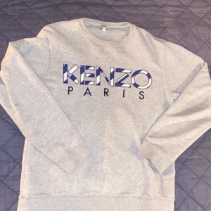 Grå sweatshirt från Kenzo - Säljer en grå sweatshirt från Kenzo med stort tryck 'KENZO PARIS' i blått och vitt framtill. Tröjan har rund halsringning och långa ärmar. Perfekt för en avslappnad och stilren look.