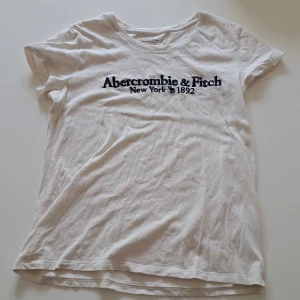 Vit t-shirt från Abercrombie & Fitch - Vit t-shirt från Abercrombie & Fitch med svart tryck på bröstet där det står 'Abercrombie & Fitch New York 1892'. Klassisk rund halsringning och korta ärmar. Tillverkad i 100% bomull för en mjuk och skön känsla. Skriv gärna vid funderingar🙂