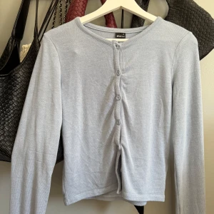 Ljusgrå kofta från Gina Tricot - Säljer en ljusblå kofta från Gina Tricot med rund halsringning och knäppning framtill. Tröjan har långa ärmar och är tillverkad i ett mjukt, tunt material som passar perfekt till lager-på-lager.