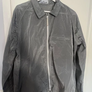 Grå overshirt från Stone Island - Säljer en grå Stone Island overshirt med dragkedja i perfekt skick, knappt använd. Köptes under en tuff period när jag var överviktig – den gav mig trygghet och stil. Nu, efter en stor viktnedgång, passar den inte längre. Det är med blandade känslor jag säljer den, men hoppas den får betyda lika mycket för någon annan