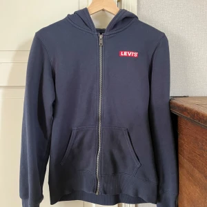 Mörkblå hoodie från Levi's - Säljer en mörkblå hoodie från Levi's med dragkedja och huva. Tröjan har två fickor framtill och en röd Levi's-logga på bröstet. Perfekt för en avslappnad stil och enkel att matcha med det mesta.