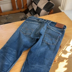 Replay Anbass jeans blå tapered - Säljer ett par snygga Replay Anbass jeans i klassisk blå tvätt med coola slitningar och detaljer. Modellen har tapered passform, fem fickor och logotyp vid linningen. Jeansen är gjorda i mjukt denim och har normal midja. Perfekta för dig som gillar stilrena och bekväma jeans.