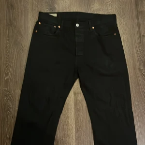 Svarta Levi's 501 jeans W33 L32 - Svarta Levi's 501 jeans med klassisk rak passform och knäppning framtill. Byxorna har fem fickor, nitar i kopparfärg och den ikoniska läderpatchen bak i midjan. Tillverkade i slitstarkt jeanstyg och passar perfekt till en avkopplad look.