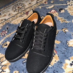 Svarta sneakers med röd sula Louboutin - Snygga svarta sneakers från Christian Louboutin med klassisk röd sula och svarta skosnören. Ovandel i textil och detaljer i skinn, rund tå och platt sula. Perfekta för dig som vill sticka ut med en ikonisk detalj under skon.Storlek 41 och levereras med låda.Skorna är helt nya och har endast använts en gång.
