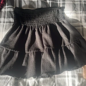 Svart volangkjol från Gina Tricot - Supersöt svart kortkjol från Gina Tricot med smockad midja och volanger. Kjolen har en flowy känsla och är perfekt för dig som gillar en feminin och trendig stil. Enkel att matcha med både crop tops och stickade tröjor.