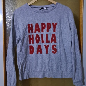 Grå sweatshirt med röd text - Grå sweatshirt med stora röda bokstäver på bröstet som stavar 'HAPPY HOLLA DAYS'. Tröjan har rund halsringning och långa ärmar. Tillverkad i mjuk bomullsmix, perfekt för en chill och avslappnad stil.