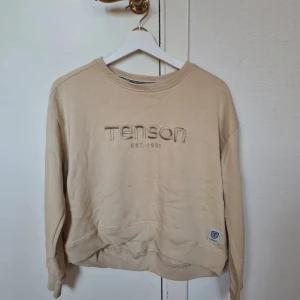 Beige sweatshirt från Tenson - Beige sweatshirt från Tenson med broderad logga och text på bröstet. Tröjan har rund hals, ribbade muddar och är tillverkad i mjuk bomull. Perfekt för en chill och avslappnad stil. Långärmad modell med klassisk passform. Liten fläckar på framsidan som går bort I tvätten 
