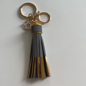 Grå och guldig väsk reflex  - Snygg väskdekoration med tofs i grått och guld, perfekt att hänga på din favoritväska eller nyckelknippa. Har guldfärgade metalldetaljer och en liten blomma som extra detalj. Ger en lyxig och trendig touch till din look.