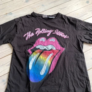 Rolling Stones t-shirt från Zara - Svart t-shirt från Zara med ikoniskt Rolling Stones-tryck framtill. Stor färgglad tunga i rosa, blått, gult och vitt. Klassisk rund hals och korta ärmar. Perfekt för dig som vill ha en cool och rockig vibe. Materialet är mjuk bomull.