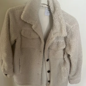 Beige teddyjacka från Lindex Kids - Supermjuk och fluffig teddyjacka från Lindex i beige färg. Jackan har stora fickor framtill, knappar och en bred krage. Perfekt för dig som vill ha en mysig och trendig look under kyliga dagar. Använd 2-3 gånger.