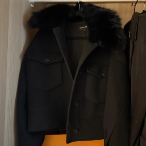 Svart ulljacka med fuskpäls från Meotine - Säljer en svart, croppad ulljacka från Meotine med stora bröstfickor och knappar framtill. Jackan har en fet krage i fuskpäls som ger en lyxig vibe. Perfekt för dig som vill sticka ut i höst och vinter. Passar dig som gillar trendiga och stilrena plagg.