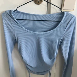 Ljusblå ribbad långärmad topp - Supersnygg ljusblå långärmad topp med ribbad struktur och rundad halsringning. Toppen har snörning i sidorna för en extra detalj och är croppad i modellen. Perfekt att styla med jeans eller kjol för en fräsch look.