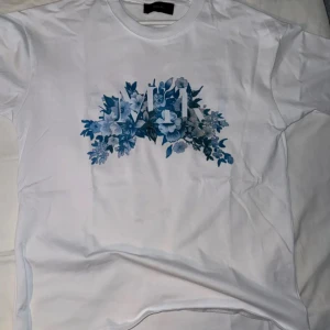Vit t-shirt från Amiri - Snygg vit t-shirt från Amiri med blått blommigt tryck och logga på bröstet. Klassisk rund hals och korta ärmar. Perfekt för dig som gillar stilrena plagg med coola detaljer. Materialet är mjuk bomull som känns skönt mot huden. Den är medium men lite stor i storleken 