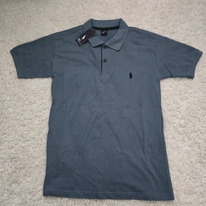 Blå pikétröja från Polo Ralph Lauren - Snygg blå pikétröja från Polo Ralph Lauren i klassisk modell med krage och knappar framtill. Tröjan har korta ärmar och den ikoniska broderade loggan på bröstet. Perfekt för en clean och stilren look.