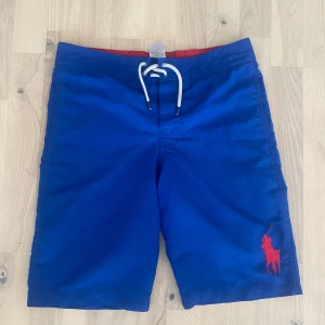 Blå badshorts från Polo Ralph Lauren - Snygga blå badshorts från Polo Ralph Lauren med röd broderad logga på benet och vit snörning i midjan. De står de är L i barn fast jag är själv S/M och säljer de för de är för stora för mig. 