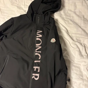 Svart vindjacka från Moncler med huva - Svart vindjacka från Moncler med stor grå logga längs framsidan och klassisk patch på ärmen. Jackan har huva, dragkedjor på fickor och bröst, samt justerbar nederkant. Väldigt bra kvalitet bara använd ett par gånger.