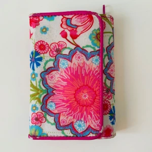 Blommig plånbok i bomull - Färgglad plånbok med blommigt mönster i rosa, blått och grönt. Plånboken har flera fack för kort, myntfack med dragkedja och en transparent ficka. Tillverkad i mjuk bomull med rosa kantband och tryckknapp. Lite skav enligt bild 