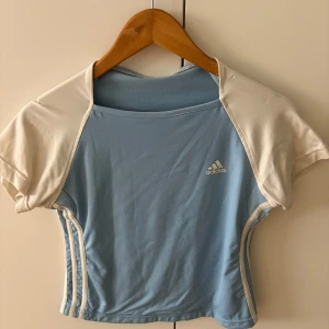 Vintage Adidas ljusblå croppad topp - Sportig ljusblå croppad topp från Adidas med vita korta raglanärmar och klassiska vita ränder längs sidorna. T-shirten har Adidas-logga på bröstet och är gjord i mjuk polyester som andas. Perfekt för dig som gillar en clean och fräsch look.