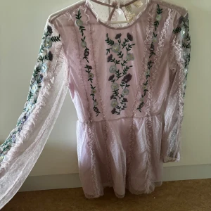 Ljusrosa broderad blommig spetsklänning  - Superfin ljusrosa kortklänning från HM med transparent spets, broderade blommor i lila, ljusblått och vitt, volangdetaljer och öppen rygg. Långärmad och med prickigt mönster i tyget. Perfekt för dig som gillar romantisk och söt stil. 