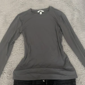 Grå långärmad topp från H&M - En stilren grå långärmad topp från H&M i storlek S. Modellen är figurnära med rund halsringning och slät yta. Perfekt att styla med jeans eller kjol för en clean look. 