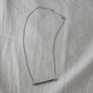 Silvrigt halsband med minimalistisk stav - Ett stilrent halsband i silverfärg med en tunn kedja och en rak, avlång stav som hänge. Perfekt för dig som gillar enkel och modern design. Lätt att matcha med andra smycken och passar till alla tillfällen.