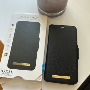 Svart plånboksfodral iDeal of Sweden - Snyggt svart plånboksfodral för iPhone 11 Pro Max/XS Max från iDeal of Sweden. Fodralet har två kortfack, magnetstängning och guldfärgad logga. Tillverkat i strukturerad syntet med stilren look och plats för både mobil och kort.
