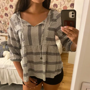 Mönstrad blus från House of Lola XS - Superskön blus från House of Lola i storlek XS. Blusen är vit med svart grafiskt mönster, har trekvartsärm och snygg snörning vid halsen. Lätt och luftig modell som sitter avslappnat och passar perfekt till jeans eller shorts.