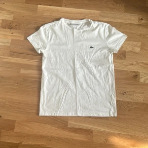 Vit t-shirt från Lacoste  - Klassisk vit t-shirt från Lacoste i 100% pima bomull. Regular fit med korta ärmar och den ikoniska gröna krokodilloggan broderad på bröstet. Enkel och stilren design som passar perfekt till jeans eller shorts.