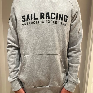 Grå hoodie från Sail Racing - Snygg ljusgrå hoodie från Sail Racing med svart tryck framtill där det står 'Sail Racing Antarctica Expedition'. Tröjan har huva, känguruficka och långa ärmar. Perfekt för dig som gillar en clean och sportig stil.