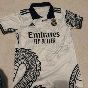 Real Madrid draktröja Adidas vit - Säljer en Real Madrid fotbollströja från Adidas i vit med svart drakmönster och marinblå detaljer. Tröjan har korta ärmar, krage och tryck med Emirates Fly Better på bröstet. Materialet är lätt och andas, perfekt för träning eller match.